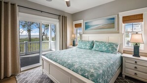 A bedroom with a large bed, a ceiling fan, a nightstand with a lamp, a window with a view of the outdoors, and a painting on the wall.