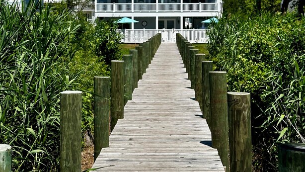 A wooden boardwalk leading to a large, multi-story building with balconies, surrounded by greenery.