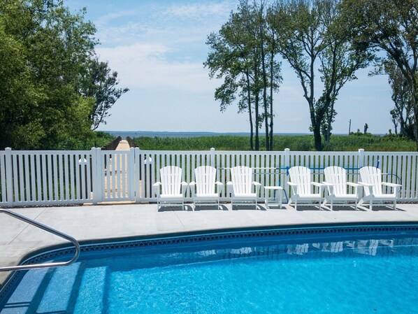 A swimming pool with white chairs and a white fence, surrounded by trees and a view of the ocean. - SOUNDFRONT with Private Pier! Private Pool, Dogs ok! (Corolla)