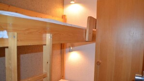 Una cama litera con estructura de madera y una cama individual debajo. Hay una puerta a la derecha y una lámpara sobre la cama.