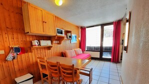 Una habitación compacta con interior de madera, una mesa de comedor con sillas, un pequeño rincón de cocina con microondas y un amplio ventanal con cortinas.