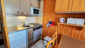 Cocina compacta con lavamanos, estufa, microondas y un refrigerador pequeño. Se observan además armarios de madera y una silla de madera.