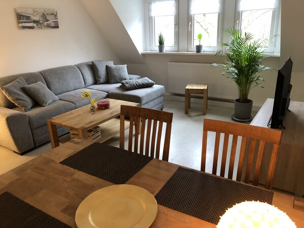 A modern living room with a grey sofa, wooden coffee table, and a dining table with chairs. There are potted plants on the windowsill and a radiator below.