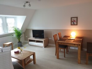 A cozy living room with a TV, wooden dining table, chairs, a potted plant, and a lamp.