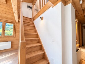A wooden staircase with a handrail, a white wall, and a window with a view of greenery.