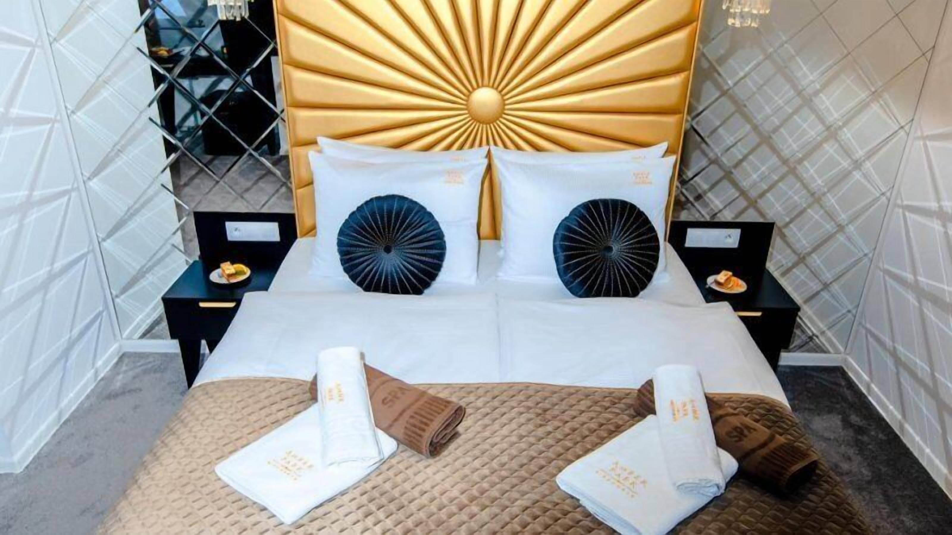 Habitación de hotel con diseño moderno, que incluye una cama grande con ropa de cama blanca, dos almohadas decorativas negras y un cabecero dorado. Hay dos mesitas de noche con lámparas pequeñas y una bandeja de aperitivos.