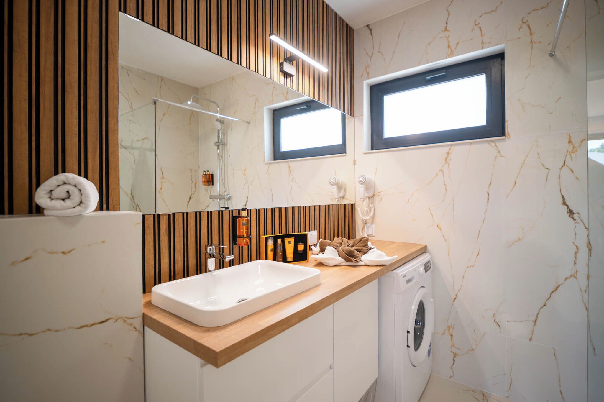 A modern bathroom with a marble sink, wooden vanity, built-in shower, and a wall-mounted window.