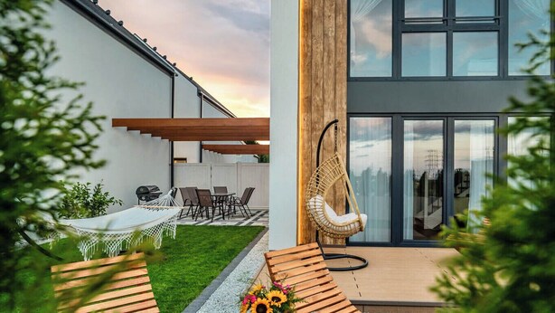 Una casa moderna con un amplio ventanal, una terraza de madera y mobiliario exterior.