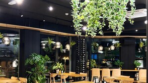 Interior de restaurante con varias mesas y sillas de madera, una lámpara con plantas colgantes y diferentes macetas.
