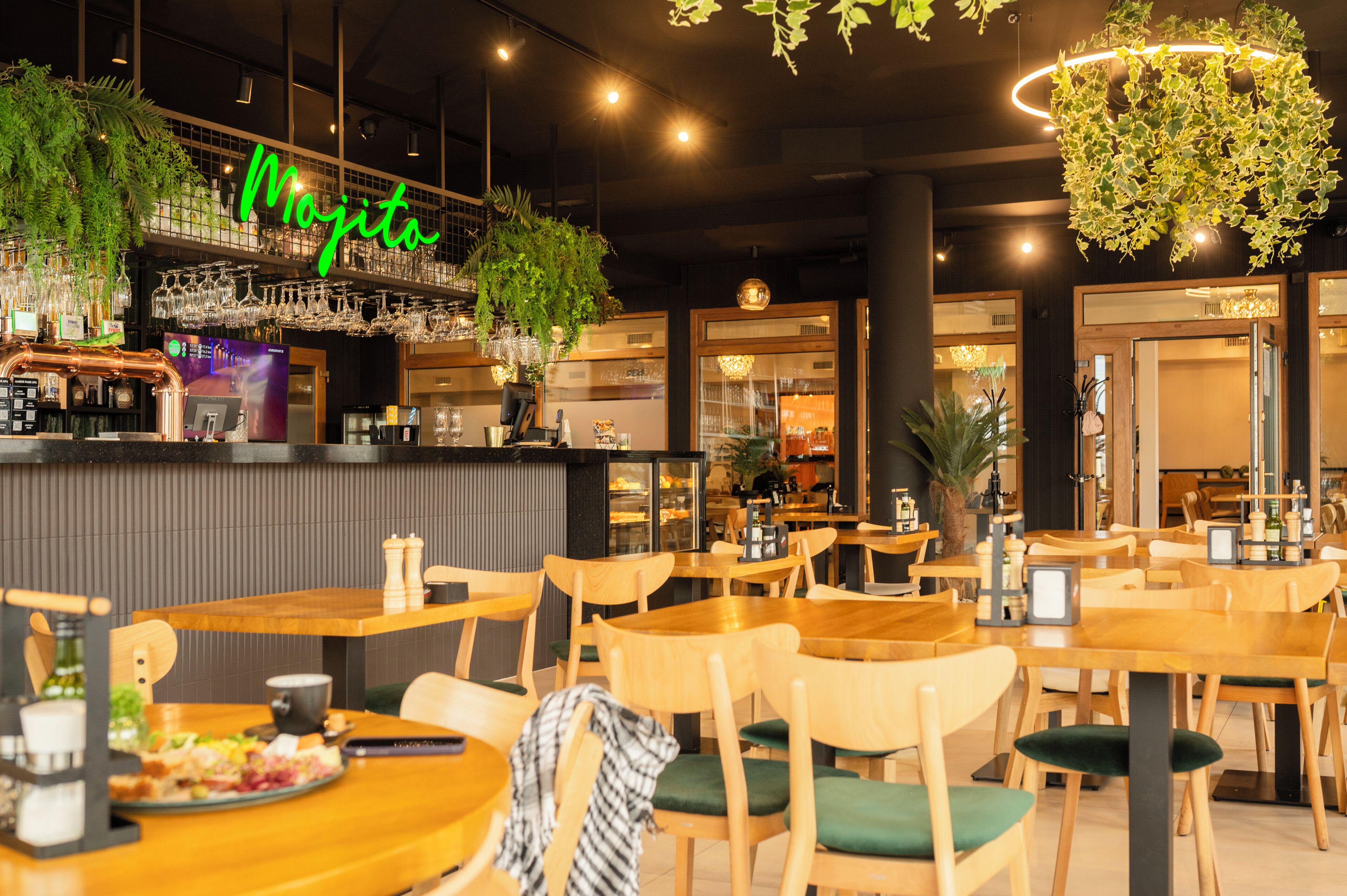 A restaurant interior with wooden tables and chairs, a bar area with a neon sign, and hanging plants.