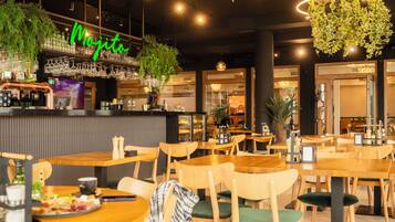 Un interior de restaurante con mesas y sillas de madera, una zona de bar con un letrero de neón y plantas colgantes.