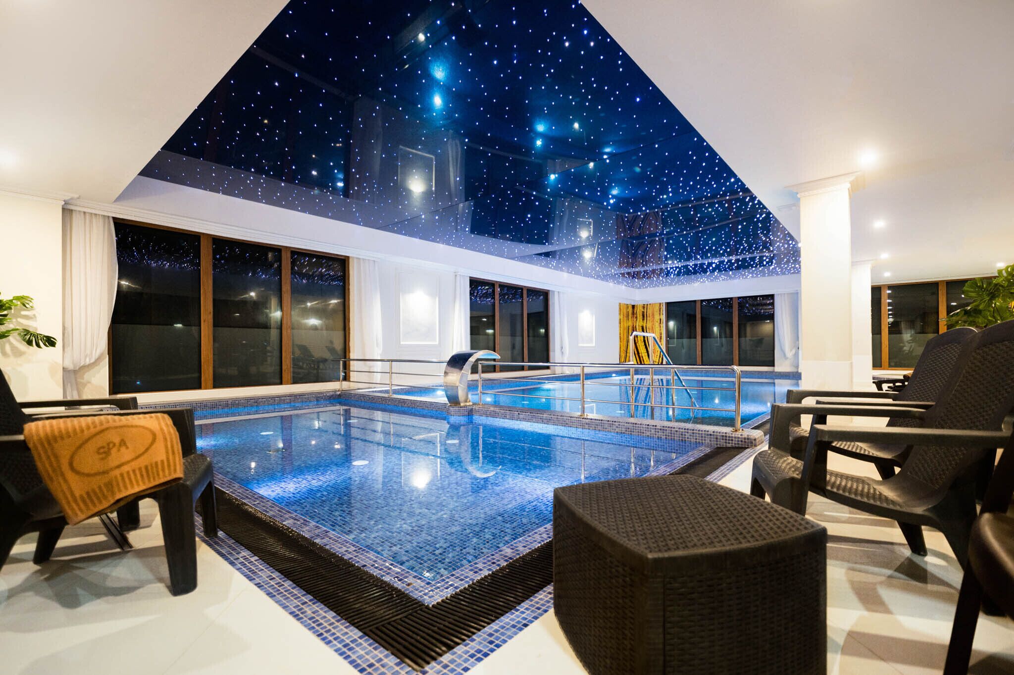 A modern indoor pool with a starry ceiling, large windows, and spa chairs.