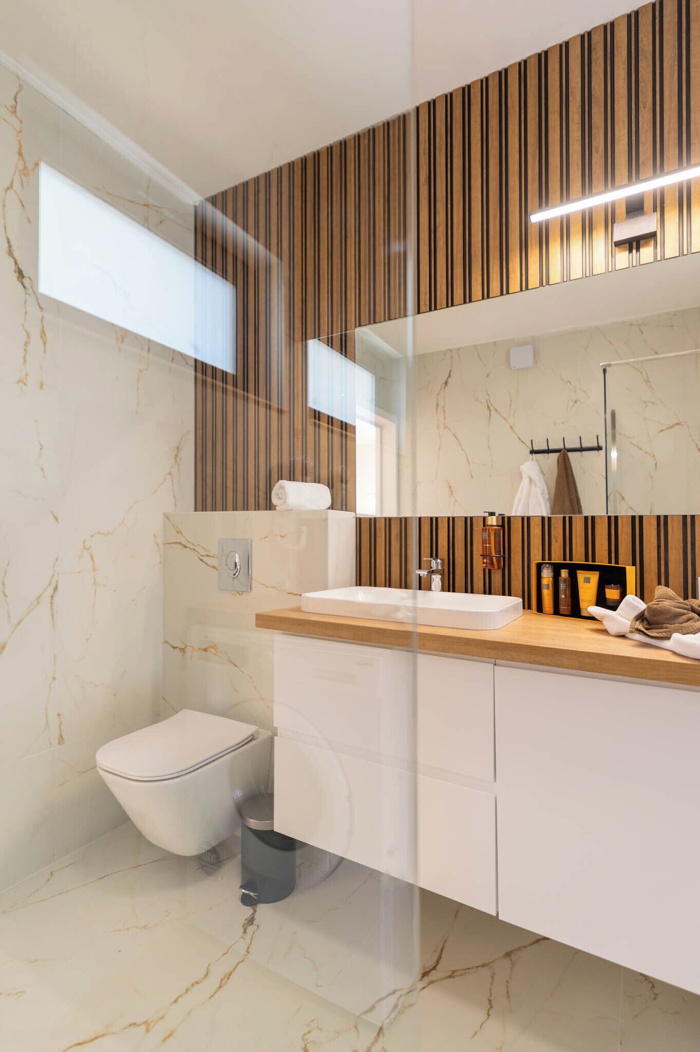 A modern bathroom with a marble floor, a wooden vanity, a white sink, a toilet, and a large mirror.