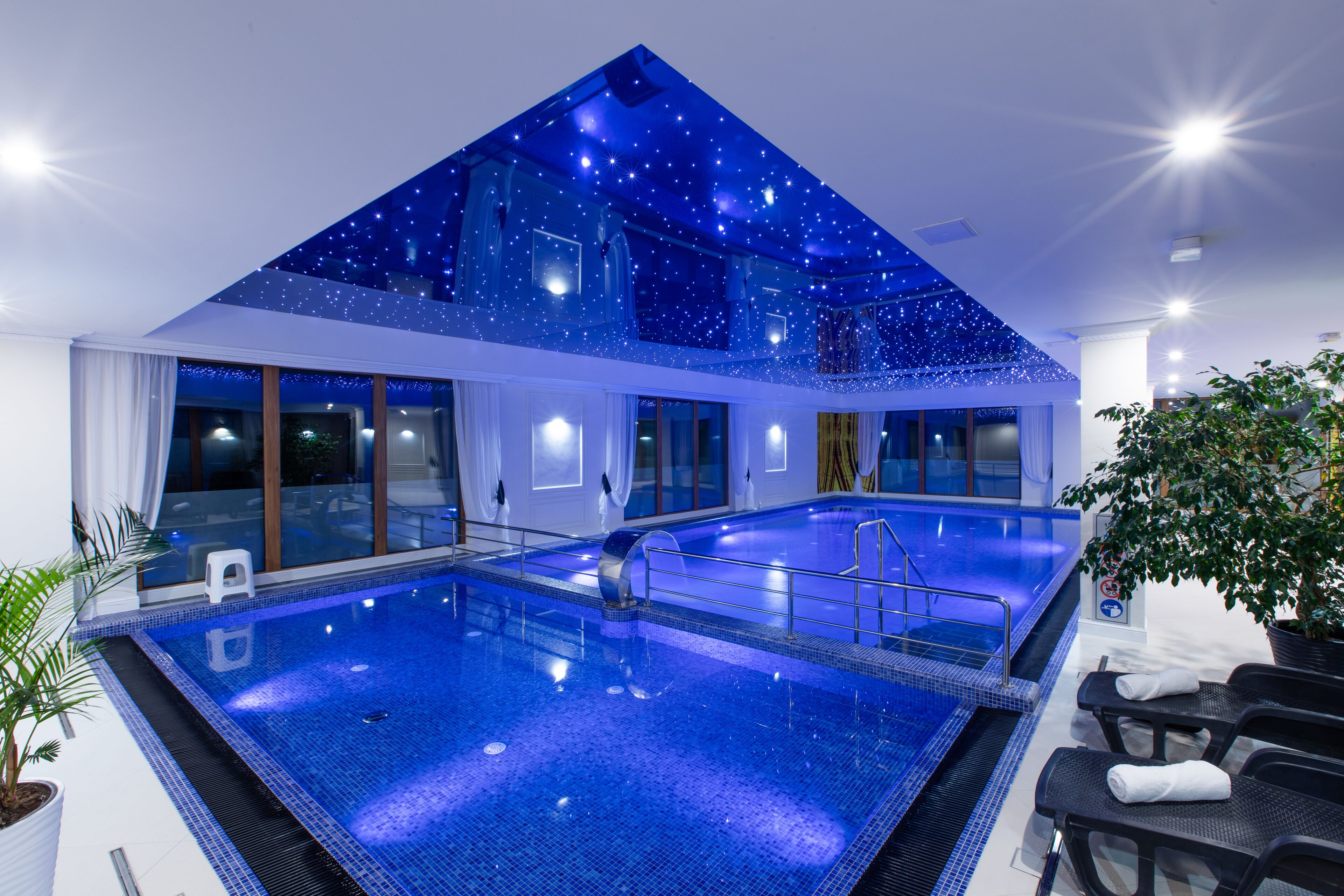 A large indoor swimming pool with a starry ceiling, surrounded by glass walls and a seating area with towels.