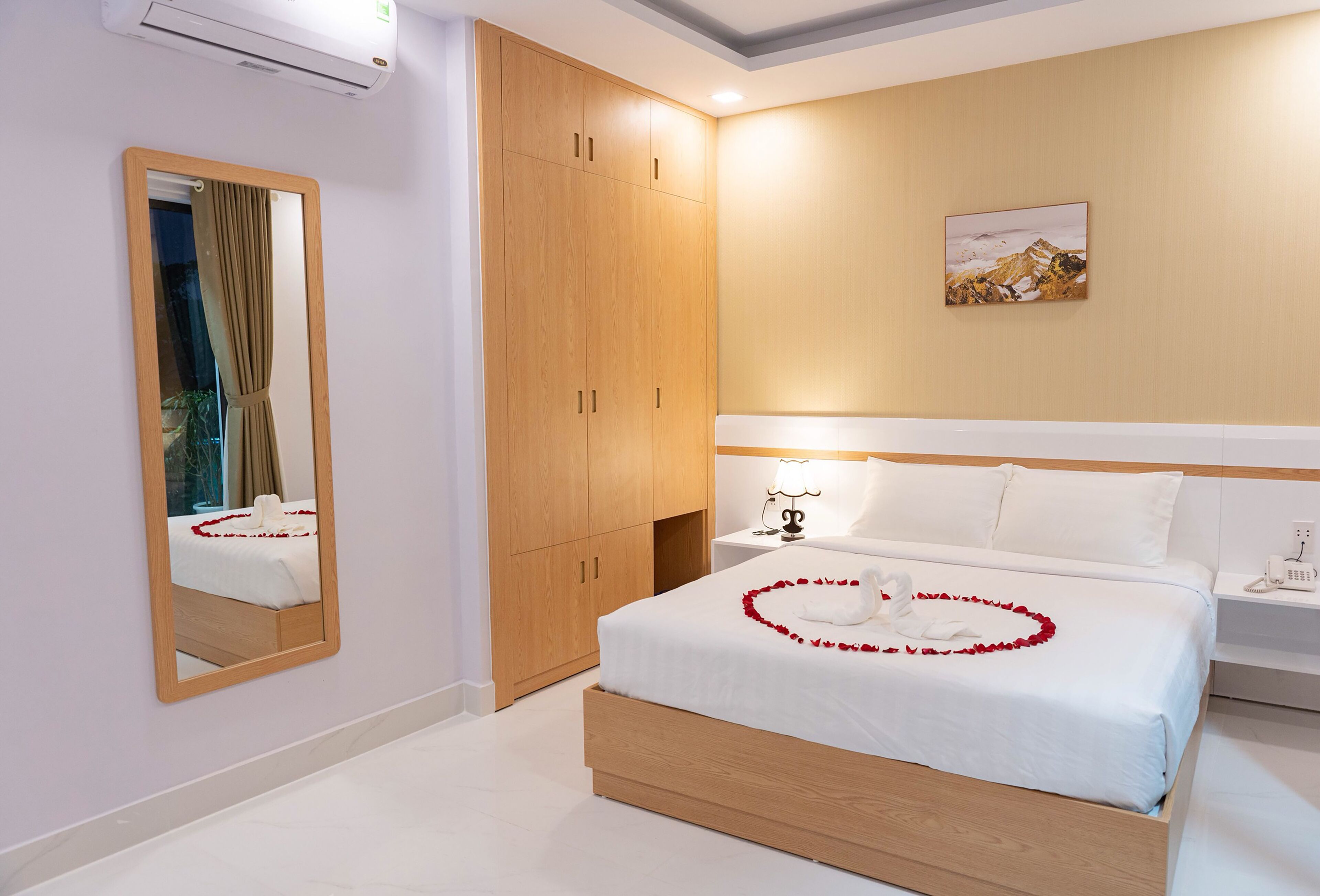 A hotel room with a large mirror, a wooden wardrobe, a neatly made bed with white bedding, and a painting on the wall.