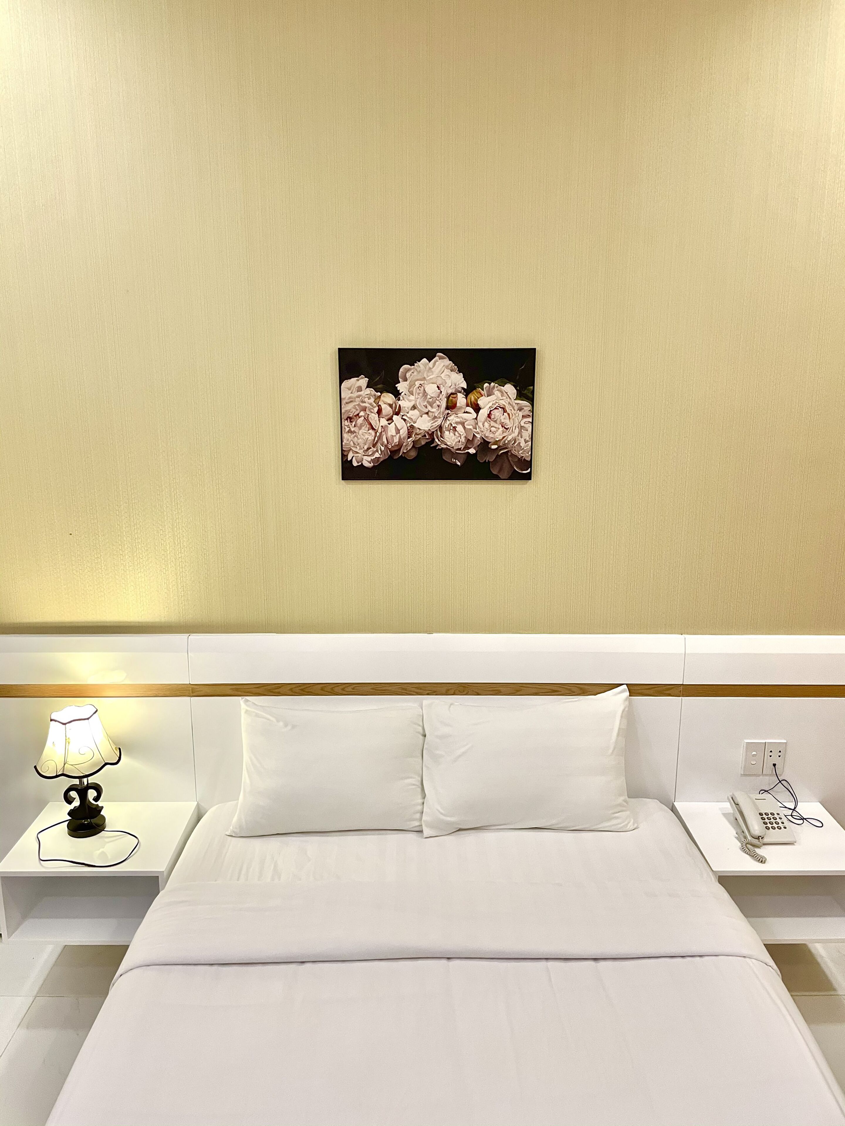 A hotel room with a bed, white bedding, a nightstand with a lamp, a phone, and a wall-mounted picture of flowers.