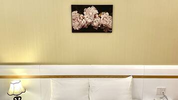 A hotel room with a bed, white bedding, a nightstand with a lamp, a phone, and a wall-mounted picture of flowers.