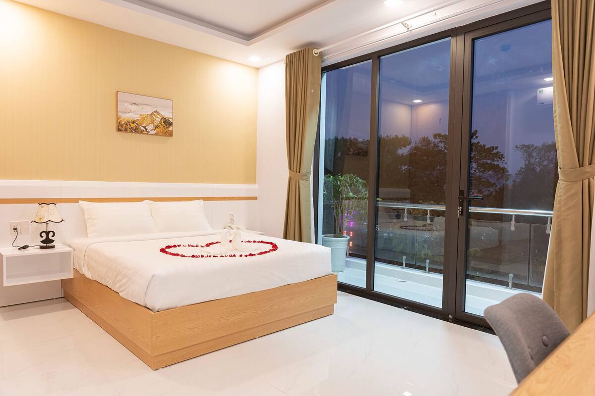 Un dormitorio moderno con una cama grande, una mesita de noche con lámpara, un cuadro enmarcado en la pared y un balcón con vistas al exterior.