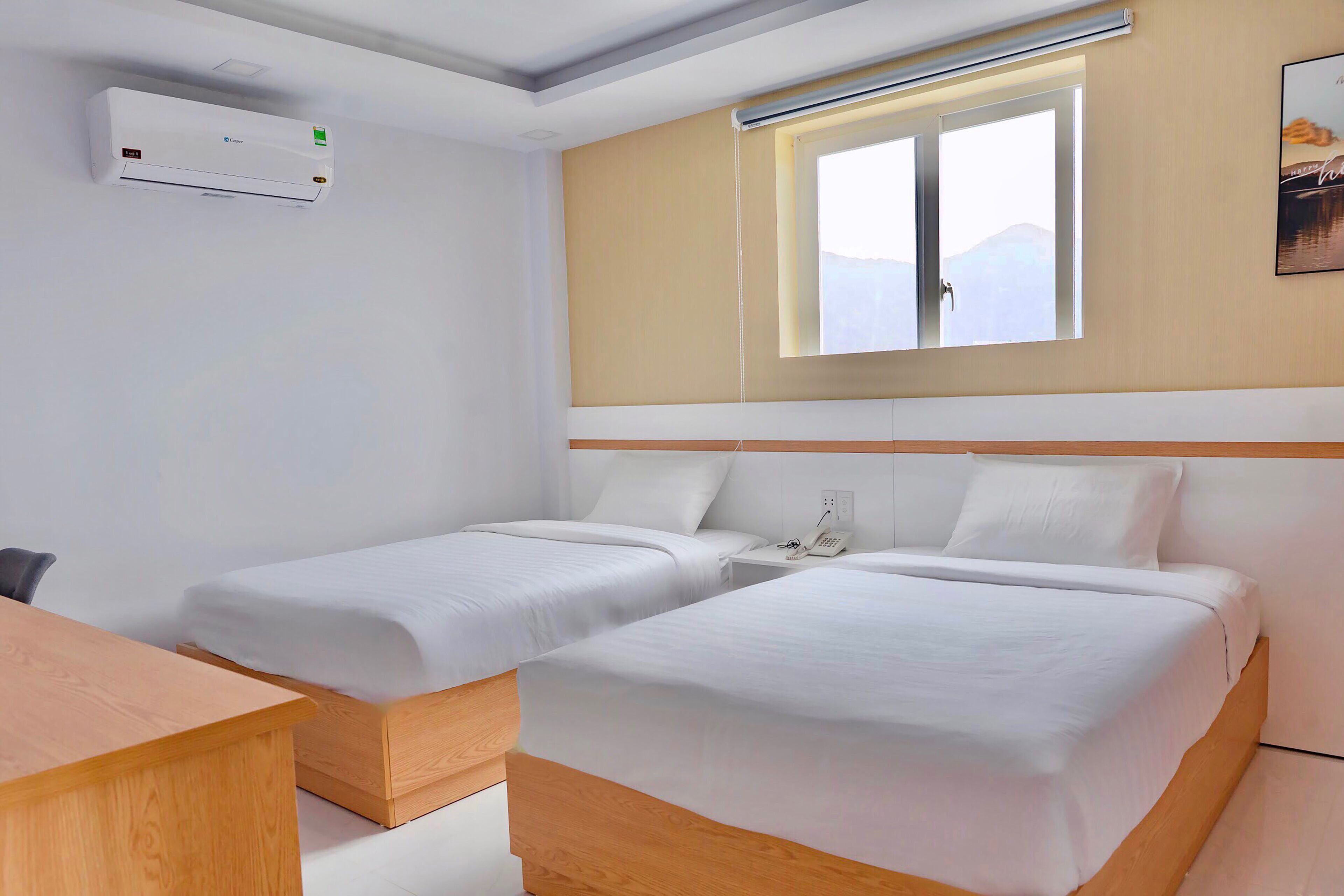A hotel room with two single beds, a desk, a chair, an air conditioning unit, a window with a view, and a wall-mounted phone.