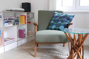 Uma sala de estar com uma poltrona verde, uma mesa de centro de vidro e uma estante repleta de livros e objetos decorativos.