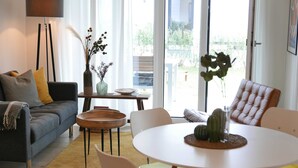 A modern living room with a sofa, chairs, a dining table, and a large window with a view of greenery. - Baabe Villen Ensemble FeWo 03 Ahoi Ref. 211406 (Baabe)