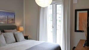 A hotel room with a bed, a nightstand with a lamp, a painting on the wall, and a view through the window. - Baabe Villen Ensemble FeWo 03 Ahoi Ref. 211406 (Baabe)