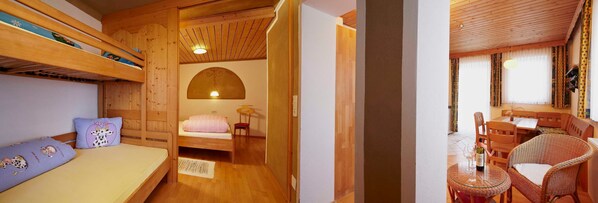 A cozy room with a bunk bed, a single bed, a wooden ceiling, and a dining area with wicker furniture.