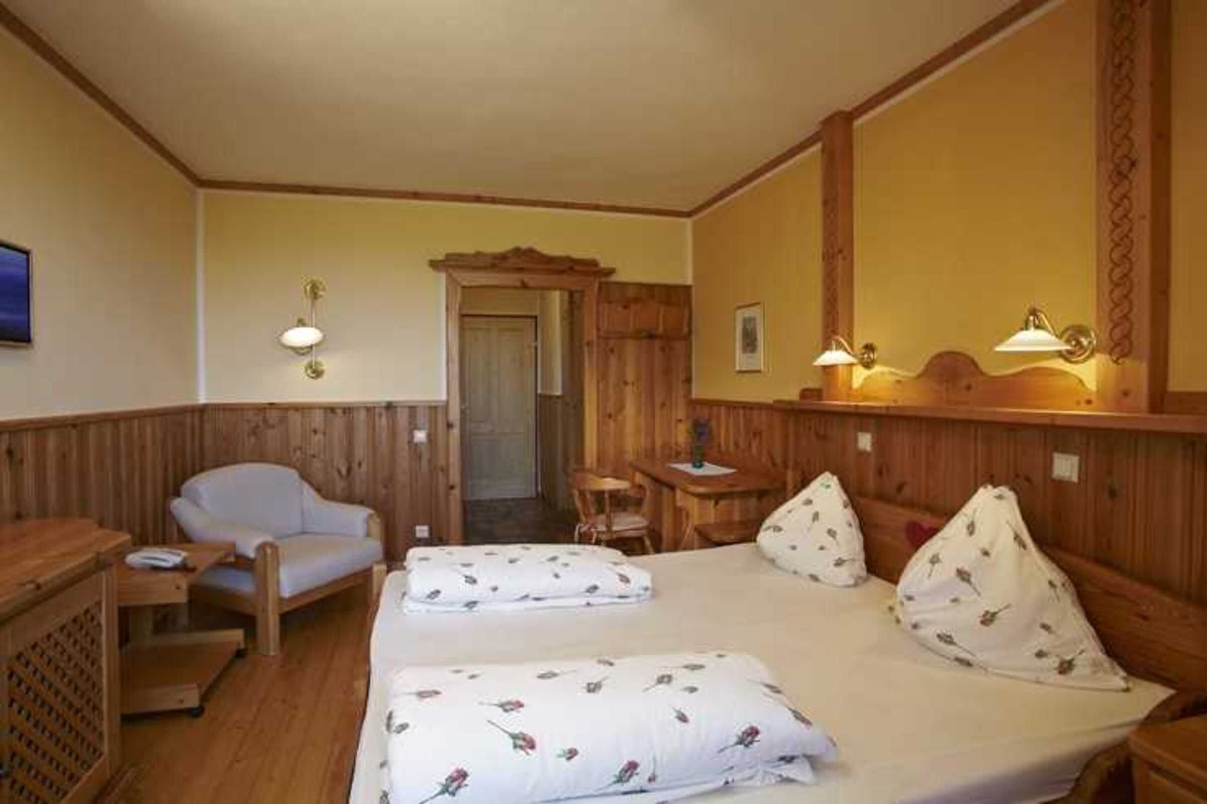 A room with wooden walls, a bed with floral bedding, a chair, a small table, and two wall-mounted lamps.