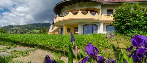 A large, curved-roofed building with a prominent balcony, surrounded by lush greenery and purple flowers.