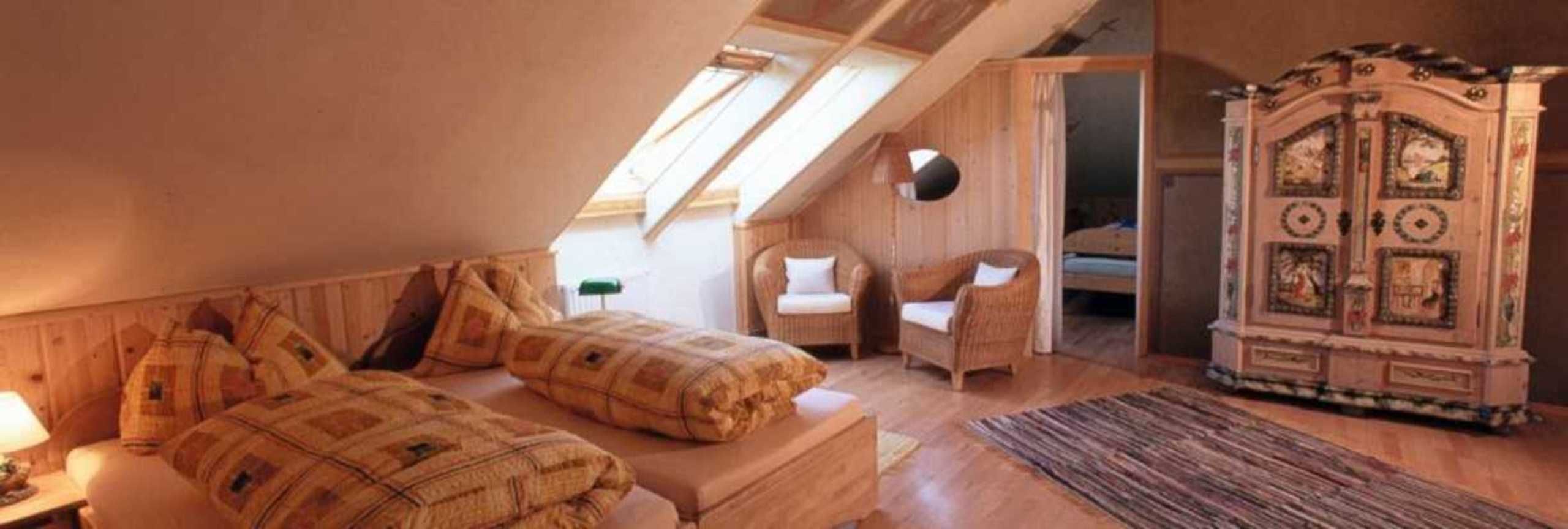 A cozy attic bedroom with a sloped ceiling, two beds with patterned bedding, a wicker chair, a cabinet with artwork, and a skylight.