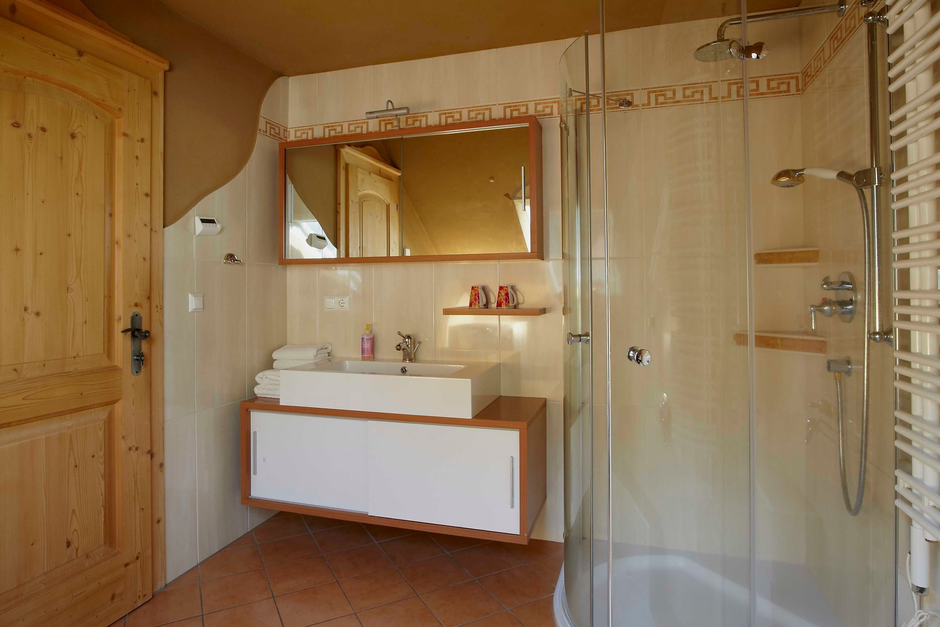 Une salle de bain avec une porte en bois, un meuble-lavabo blanc avec un miroir, une cabine de douche en verre et un sol carrelé.