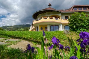 Un edificio grande con techo curvo y un balcón destacado, rodeado de exuberante vegetación y flores moradas.
