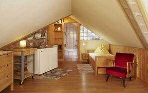 A compact attic bedroom with a kitchenette, a wooden chair, and a bed with a yellow pillow. - Bionest II earth building apartment - Biolandhaus Arche (Eberstein)