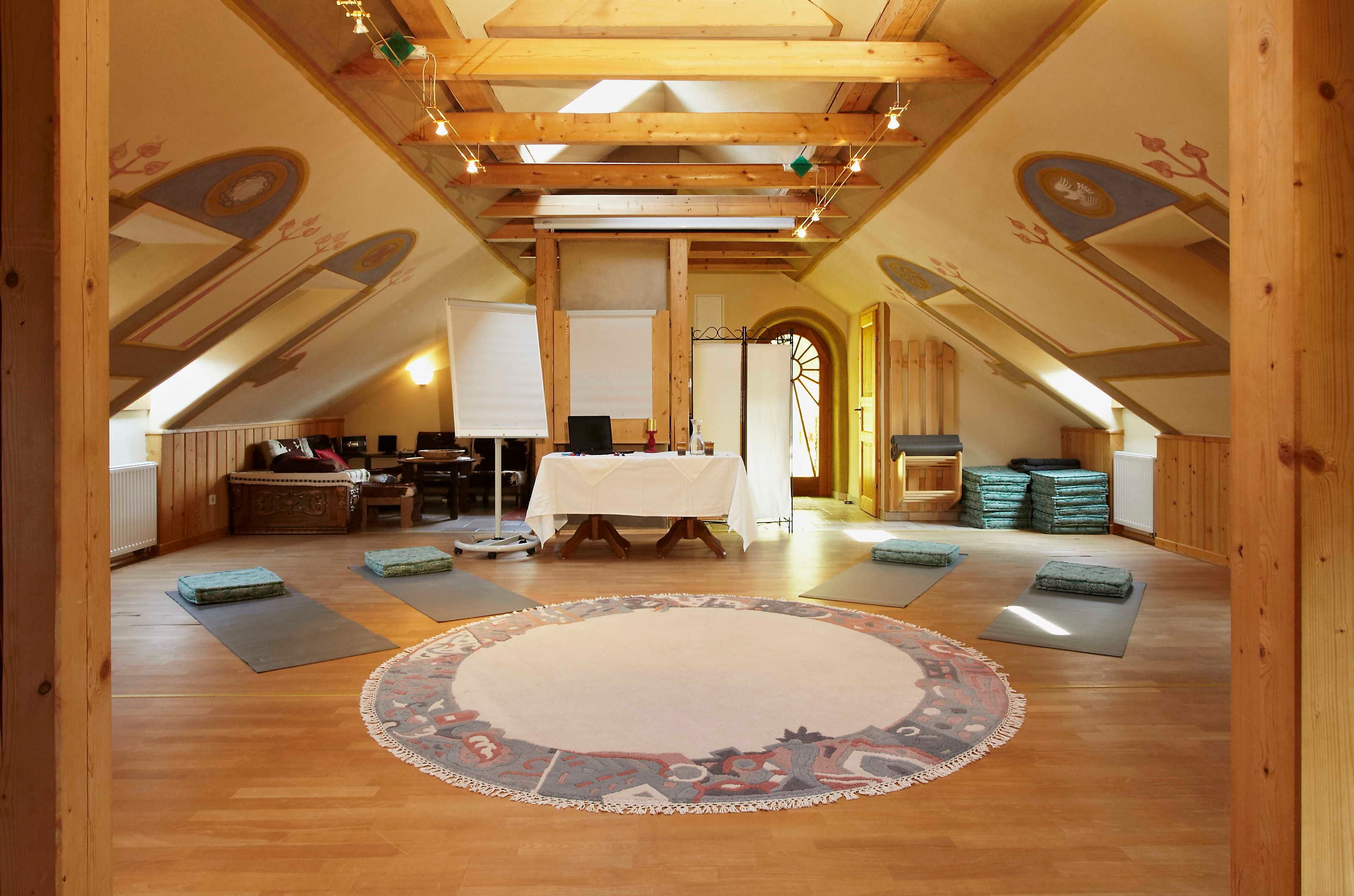 Une pièce mansardée avec un plafond haut, des poutres en bois, une table centrale recouverte d’une nappe blanche, des tapis de yoga et un tapis à motifs.