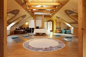 Un dormitorio en el ático con techo alto, vigas de madera, una mesa central con mantel blanco, colchonetas de yoga y una alfombra estampada.