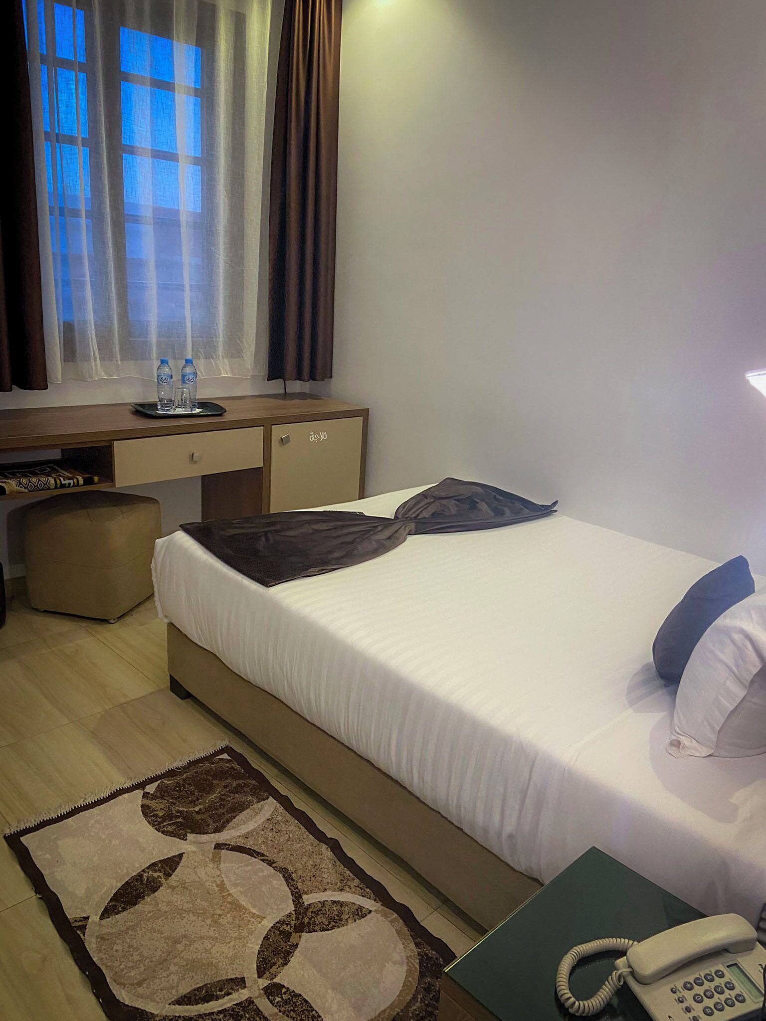 A hotel room with a bed, bedside table with a phone, a desk with a chair, a window with curtains, and a small rug.