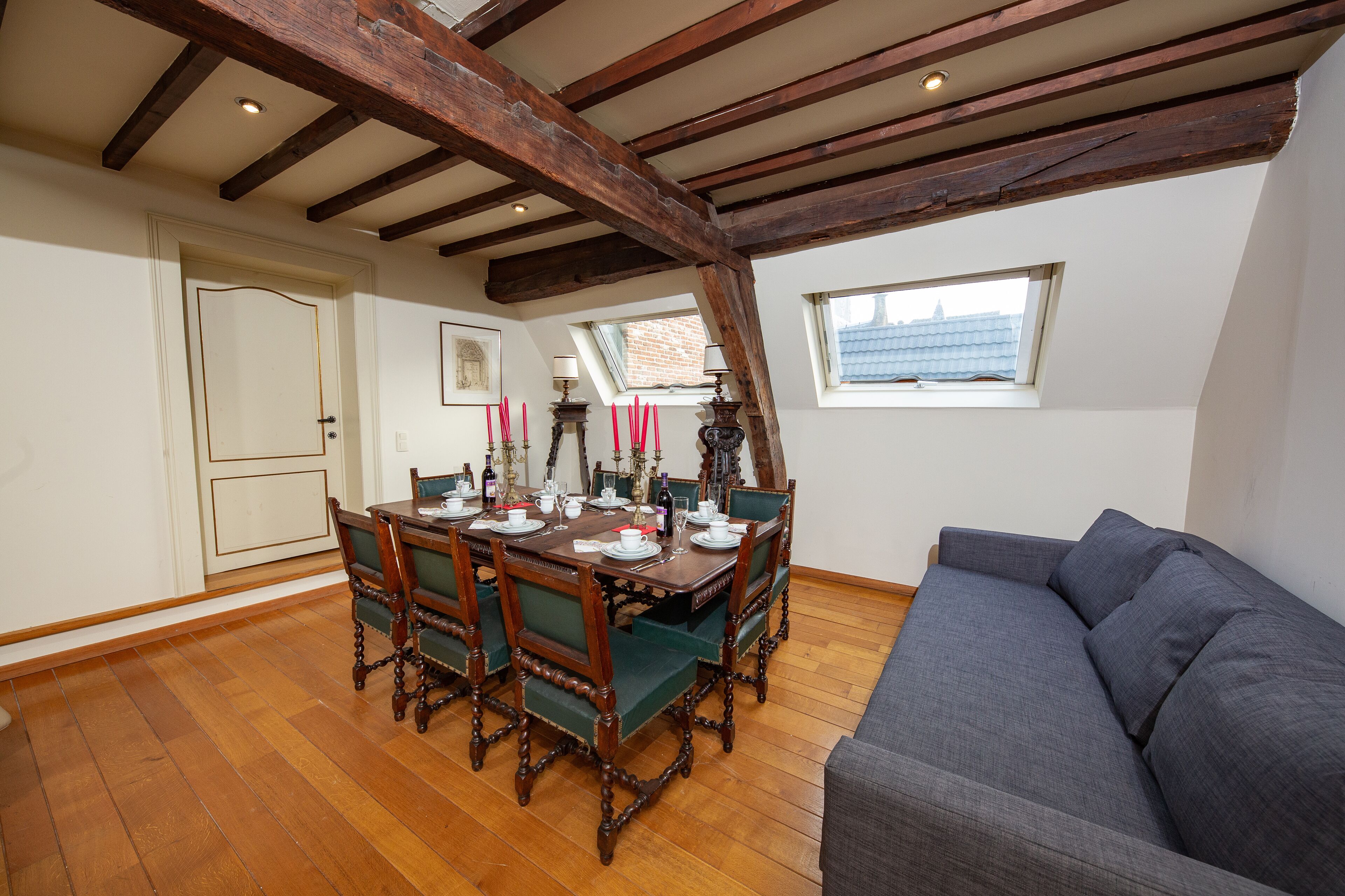 Een eetgedeelte met een houten tafel voor vier personen, omringd door groene bekleden stoelen. Een grijze bank staat rechts. De kamer heeft zichtbare houten balken in het plafond en een raam met uitzicht op gebouwen.