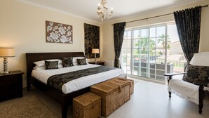 A bedroom with a large bed, a chandelier, a balcony with curtains, and a view of palm trees.
