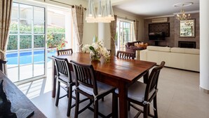 A dining area with a wooden table and chairs, a vase with flowers, and a view of a swimming pool through large windows.