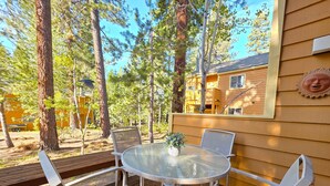 A deck with a glass table and white chairs, surrounded by trees and a wooden building. - McCloud Condo | Community Hot Tub | Deck | BBQ (Incline Village)
