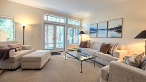 A living room with a sofa, ottoman, coffee table, and lamps. There are large windows with blinds, a door, and a wall-mounted picture. - McCloud Condo | Community Hot Tub | Deck | BBQ (Incline Village)
