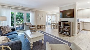 A spacious living room with a sofa, coffee table, and a TV mounted on the wall. There is a dining area with a table and chairs, and a kitchen with cabinets and a TV. - Pet Friendly | Luxury Beachwood Townhome | Directly Across the Street from the Beach (Fernandina Beach)
