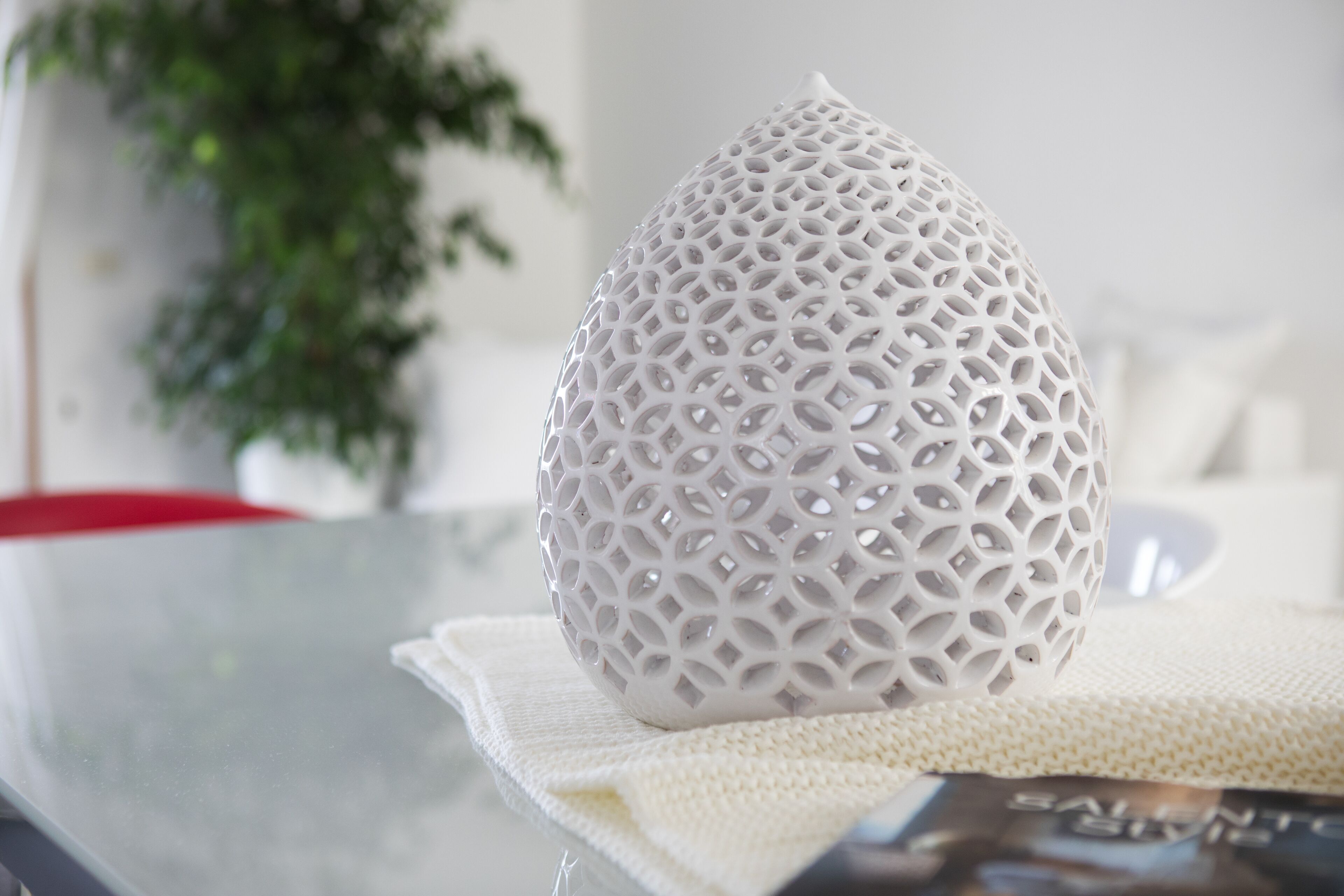 A white, intricately patterned object on a table with a white cloth, a magazine, and a green plant in the background.