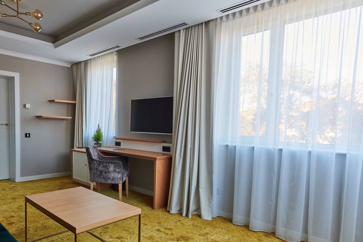 Una camera d'albergo moderna con una scrivania in legno, una televisione, una sedia e un'ampia finestra con tende leggere.
