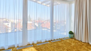 Una habitación con ventanas amplias cubiertas por cortinas blancas traslúcidas, un radiador en el suelo y una planta en maceta en el alféizar.