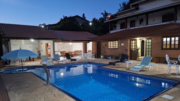 A swimming pool with lounge chairs and umbrellas, surrounded by stone tiles and a brick building with a tiled roof. - Holiday Home in Atibaia  (Atibaia)