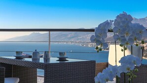 A balcony with wicker furniture, a vase of white orchids, and a view of the sea and mountains.