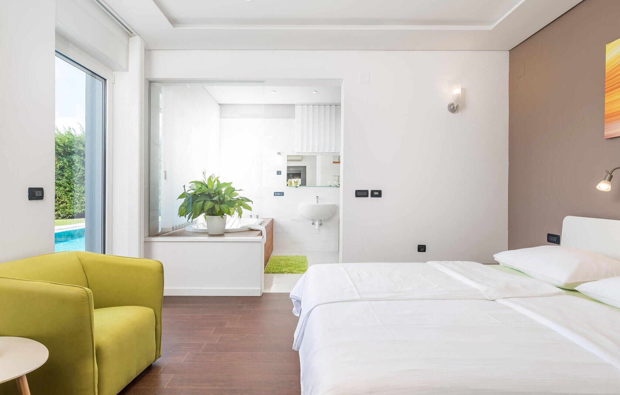 A modern hotel room with a bed, a green armchair, a small table, a potted plant, a bathroom with a sink and mirror, and a view of an outdoor swimming pool.