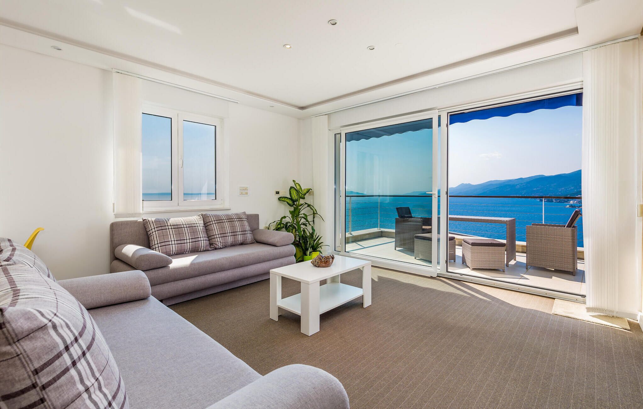 Uma sala de estar moderna com sofá, uma mesinha e vista para o mar através de amplas portas de vidro.