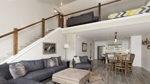 A spacious living area with a grey sectional sofa, a beige ottoman, and a wooden dining table with chairs. A staircase with wooden railings leads to an upper level. - Gorgeous Vacation Chalet: Hot Tub, Wood Stove (Big Sky)